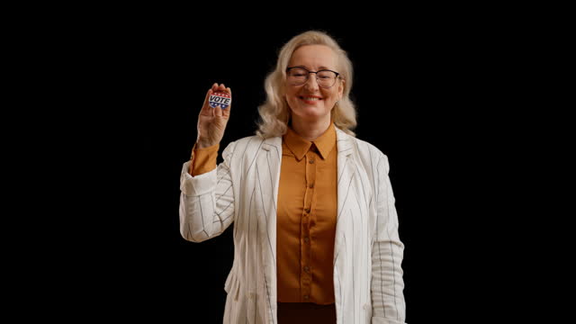 Cheerful woman in her 50s showing vote button on black background, elections