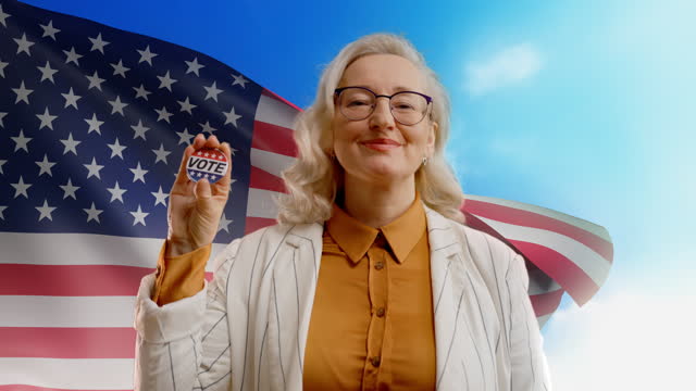 Smiling woman in her 50s showing vote button against waving USA flag, elections