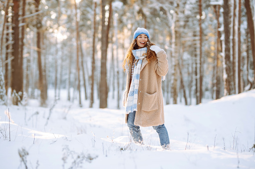 Smiling woman in a winter snow-covered park walks and plays with snow on a beautiful sunny day. Young fashionable woman in the winter forest. Walking concept, weekend.