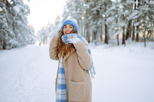 Smiling woman in a winter snow-covered park walks and plays with snow on a beautiful sunny day. Young fashionable woman in the winter forest. Walking concept, weekend.