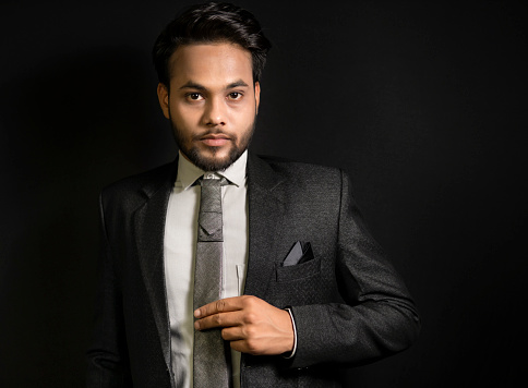 Low key side view portrait of handsome, elegant, fashionable Indian young businessman informal standing against a dark black background and looking at the camera with a blank expression.
