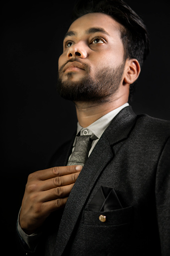 Low key side view portrait of handsome, elegant, fashionable Indian young businessman in formal dress standing against a dark black background. He is looking up with a blank expression and contemplating.