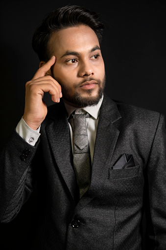 Low key side view portrait of handsome, elegant, fashionable Indian young businessman in formal dress standing against a dark black background. He is looking away with a blank expression and contemplating.