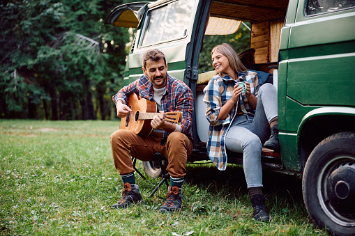 Happy man playing acoustic guitar to his girlfriend while camping in the woods.