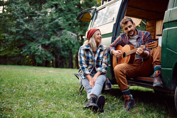 Happy camper playing acoustic guitar to his girlfriend while relaxing by their van in nature. Happy man playing acoustic guitar during camping trip with his girlfriend in the woods. Copy space. white caravans in a park stock pictures, royalty-free photos & images