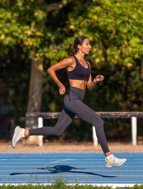 bella giovane runner abbronzata magra, concentrata, vestita con abbigliamento sportivo attillato, sospesa in aria che corre con stile e posizione perfetta del corridore su una pista blu, con stile e posizione perfetta. - jump shot foto e immagini stock