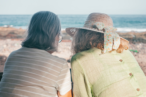 Back view of female couple of senior and middle age women with hats looking at news on mobile app. Two female friends enjoying freedom and relax in outdoors face the sea
