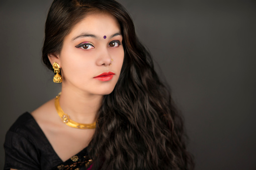 Portrait of beautiful serene Indian young woman in traditional Indian sari with gold jewelry looking at the camera with a blank expression over a dark background.
