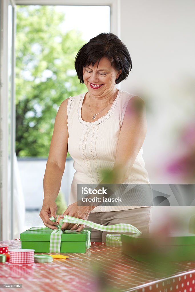 Mature woman wrapping gift Portrait of cheerful mature woman binding a ribbon on a gift box. 60-64 Years Stock Photo