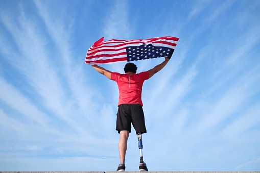 Rear view of unrecognizable young man with prosthetic leg standing holding the U.S. flag above his head with a cloudy blue sky on the background