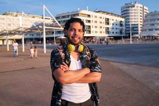 Front view of a young man standing with his arms crossed looking at the camera with his wireless headphones around his neck in the beach promenade.