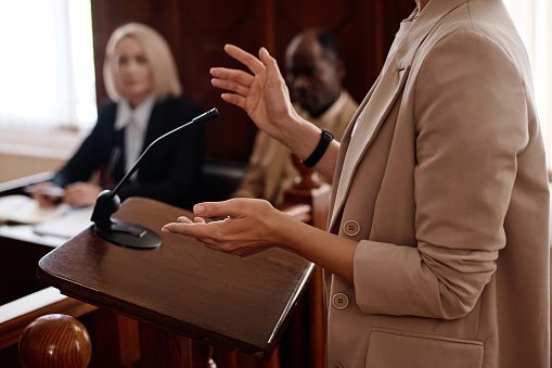 Cropped shot of young female witness in beige blazer standing by tribune with microphone and testifying in front of jury, judge and protection side