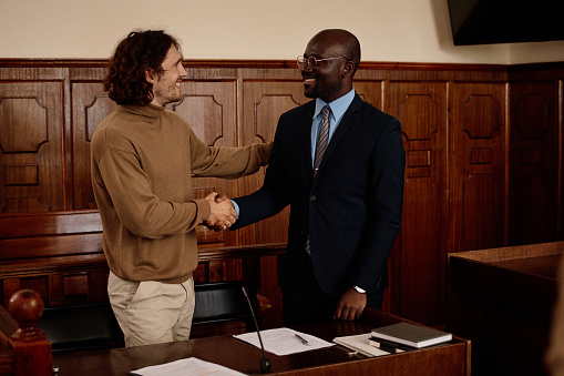 Happy young intercultural male suspect and his attorney shaking hands after announcing acquittal by judge during trial session in courtroom