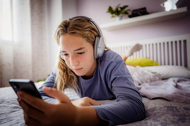 ragazza adolescente che utilizza le cuffie e la messaggistica di testo sul telefono - one teenage girl only on the phone bed lying on front foto e immagini stock