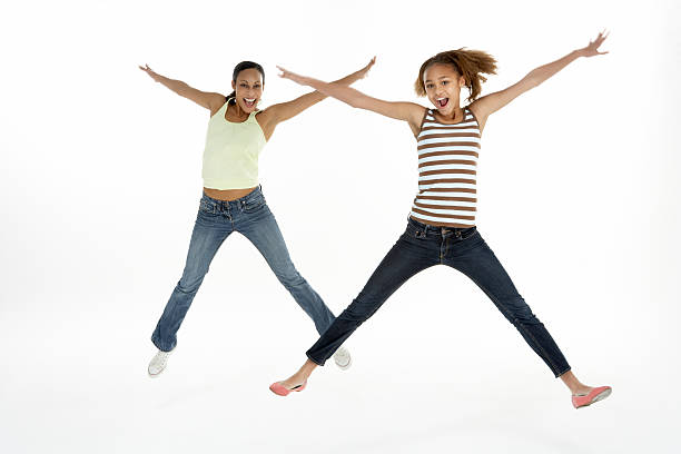 Three individuals woman, dressed in jeans and sweaters, are captured mid-jump in an outdoor field. Arms extended, they express freedom and joy. Urban buildings in the background suggest an urban park setting.