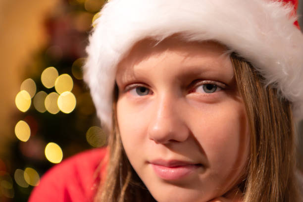 Portrait of teenage girl in Santa hat sitting on chair in front of Christmas tree, Merry Christmas and Happy Holidays!