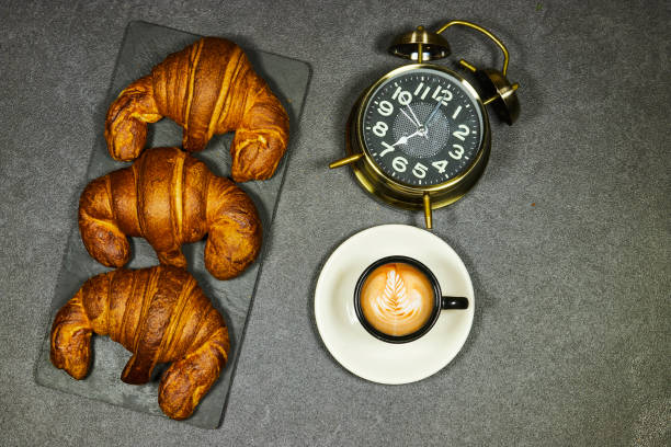 morning concept - Fresh baked croissants on black board  for breakfast and  coffee cup with leaf late art and alarm clock showing seven o'clock on gray grunge background