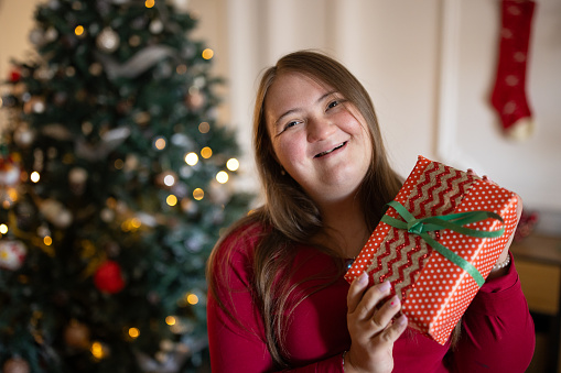 Portrait of a young Caucasian woman with Down syndrome holding a Christmas present in front of a Christmas tree in the living room of a house