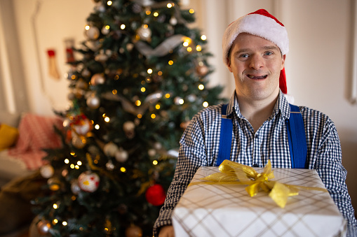 Portrait of a mid adult Caucasian man with Down syndrome holding a Christmas present in front of a Christmas tree in the living room of a house