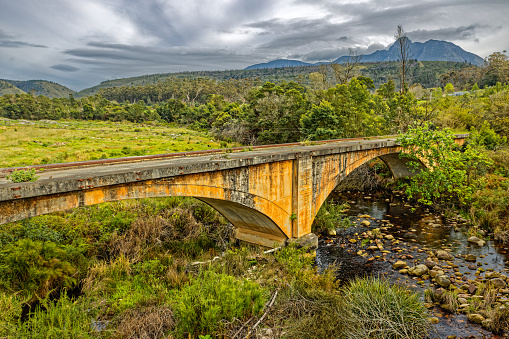 Abandoned concrete road bridge over small rocky stream with dark mountains and cloudy sky  near Riversdale, Western Cape, South Africa