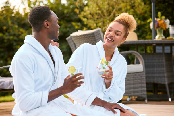 coppia che indossa accappatoi all'aperto seduta con bevande in piscina nel giorno della spa - treated water foto e immagini stock