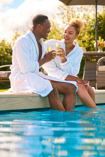 istock Couple Wearing Robes Outdoors Sitting Doing Cheers With Drinks By Swimming Pool On Spa Day 1746212587