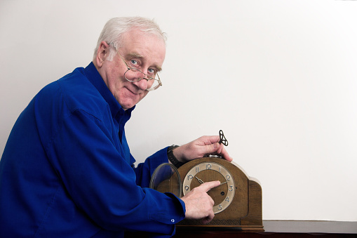 grandfather looks impishly at the camera while adjusting and winding an old fashioned Edwardian / Victorian style clock; image bleeds to white on top right hand side