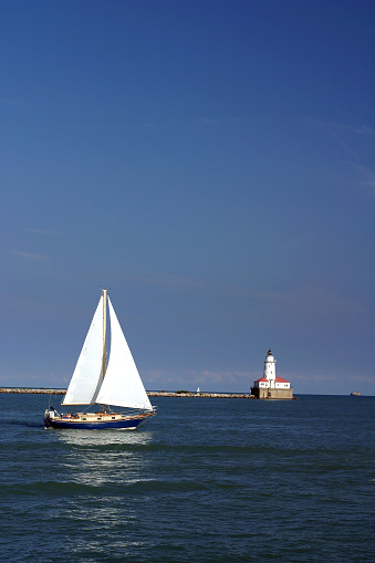 Sailboat sails by Chicago pier lighthouse with graduated blue sky.