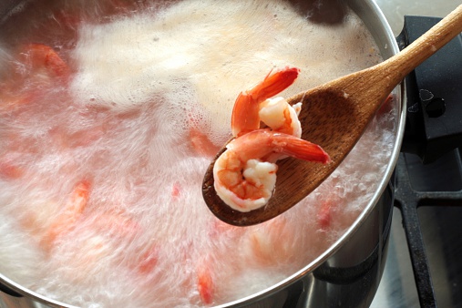 Two boiled shrimp in a wooden spoon with a pot of boiling shrimp in the background.