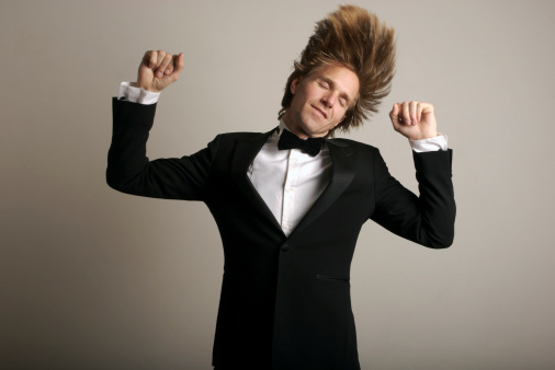 Man with long blond hair dances in tuxedo against white wall
