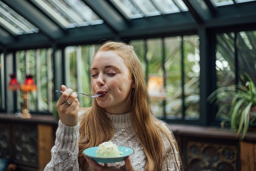 A young woman standing in a restaurant venue in Northumberland, North East England. She has her eyes closed and is enjoying a white chocolate dessert on a plate with a fork.\n\nVideos also available for this scenario.