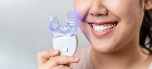The woman is whitening her teeth with a teeth whitening device by herself in the living room at home. Health and medical concept.
