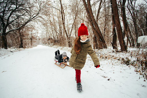 Girl pulling her brother on the sledge and having fun on the snow in the forest