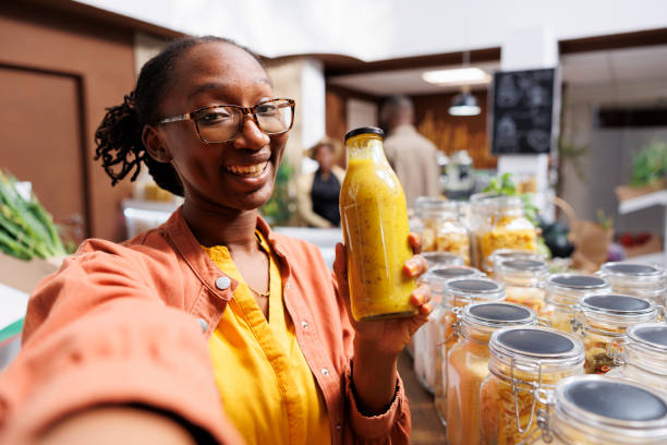 African American woman doing a video call while holding a bottle filled with pasta sauce. She uses technology for online marketing and connects with customers through vlogging.
