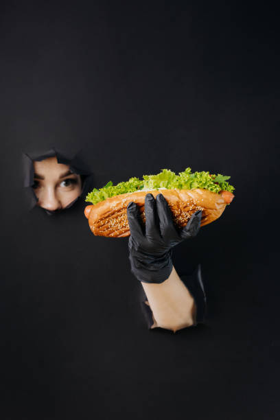 weibliche hand hält hot dog und schaut durch löcher im schwarzen papierhintergrund. streetfood-konzept - sausage grilled isolated single object stock-fotos und bilder
