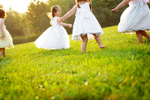 Color photo of four happy little girls wearing princess dresses and dancing in the grass together.