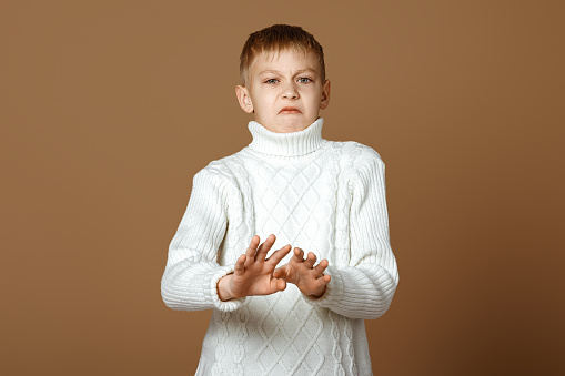 Displeased teen boy deny something, shakes hands and say no, rejects offer, looks insulted, express disapproval, stands over beige background in white knitted sweater