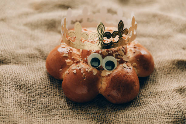 istock Three Kings Cake with a golden crown and a tiny king hidden inside. Swiss tradition to bake Dreikönigskuchen on January 6. Rustic sackcloth background, funny googly eyes. 1746384900