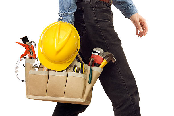 Close-up of a handyman or repairman carrying work tools, and walking with home improvement equipment including a hard hat, caulking gun, hammer, wrench, plier, screwdriver, protective eyewear safety goggles, and other hand tools for manual labor. Lower body partial view of worker wearing denim jeans, isolated on white background.