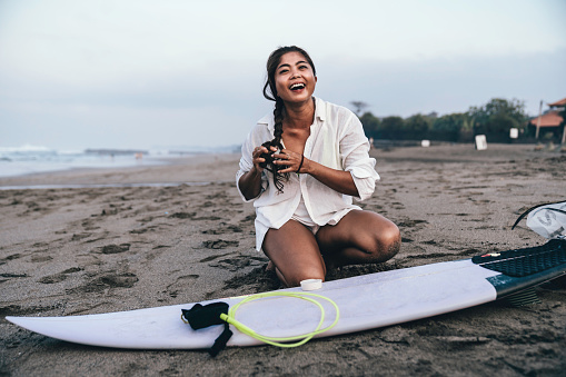 smiling woman wearing a wetsuit while kneeling on sandy beach and waxing her surfboard on the beach