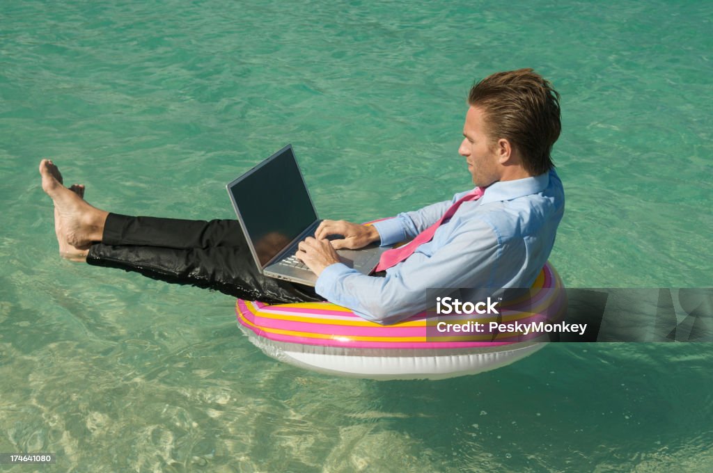 Relaxed Businessman Keeping Afloat on Tropical Sea with Laptop Businessman types on his laptop computer while floating in rubber ring in tropical blue waters Inflatable Swim Ring Stock Photo
