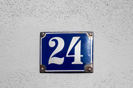 House address - number 24 on a blue enamel sign. White housewall in the background. Bavaria, Germany.