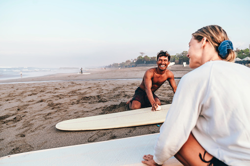 young couple kneeling on sandy beach and waxing her surfboard on the beach