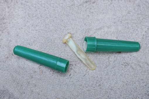 part of a brown broken pipette and an open green plastic case parts lie on the gray sand on the street