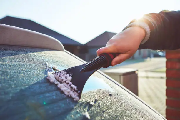 Photo of A woman scrapes frost off the front window of her car in winter