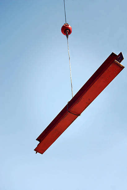 Low angle shot of a large steel I-beam being hoisted into the air with a clear blue sky background.