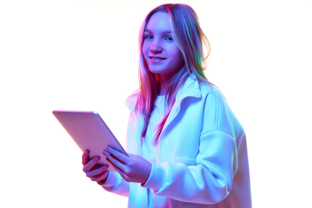 istock Smiling girl in warm winter coat in mixed neon filter holding tablet isolated white studio background. 1746574465
