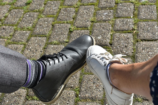 close up of 2 feet touching toes, one a mans foot wearing dark blue shoe, one woman's foot wearing white training shoe.  The people are sat down taking a rest.  Brick paver to background