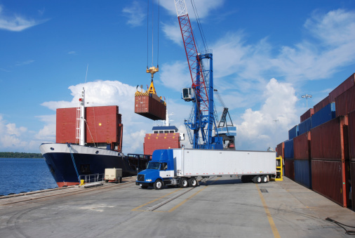 Shipping and Trucking Transportation Industry - tractor trailer sits in the foreground at a commercial dock. Containers are load on board a ship by cranes