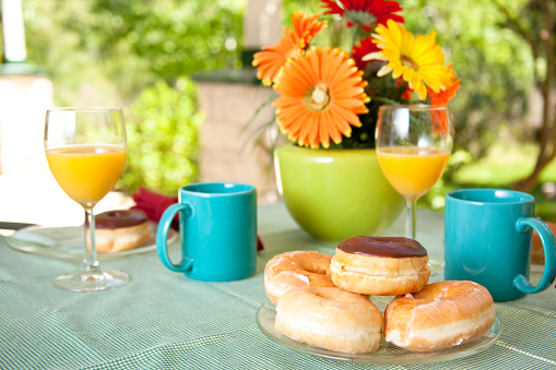 Patio table set with plate of fresh donuts and coffee.  Table cloth and flower arrangement are on table.  Yard and shrubs can be seen in the background.  See more of this series in lightbox below.
